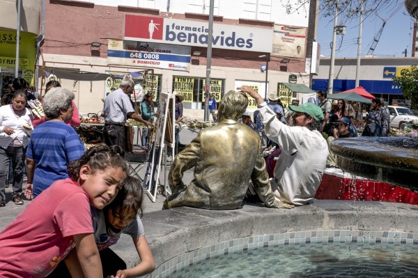 An image of a fountain in the historic center of Juarez, with a statue sitting on the rim of the fountain next to a man, and people going about their day nearby.