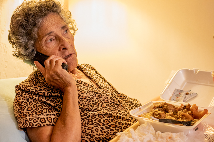 An elderly woman talks on the phone while eating Chinese food.