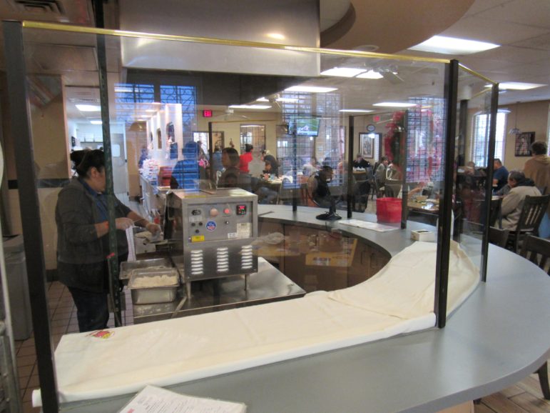 A clear divider is displayed the front of the restaurant, behind the partition a woman is making fresh tortillas.